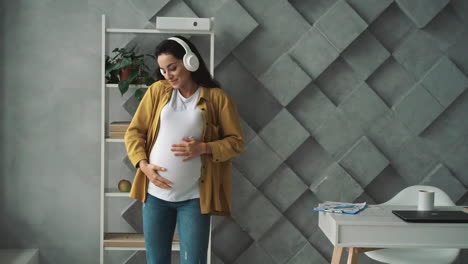 young happy pregnant woman dances listening to music in headphones while holding and stroking her belly