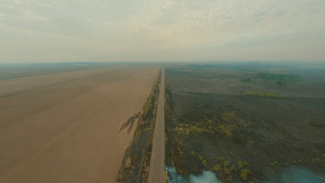 fire-next-to-road,-truck-driving-next-to-smoke-clouds,-farmland-cultivation,-burn-clearing-plantation,-drone-aerial-overview-shot,-roadside-fire