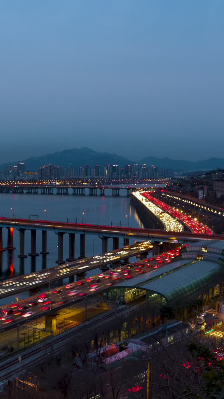 Timelapse verticale con transizione dal crepuscolo alla notte che cattura il traffico intenso dell'ora di punta sull'autostrada Gangbyeonbuk-ro e sul Ponte Dongho con i treni della metropolitana alla stazione di Oksu, Seoul