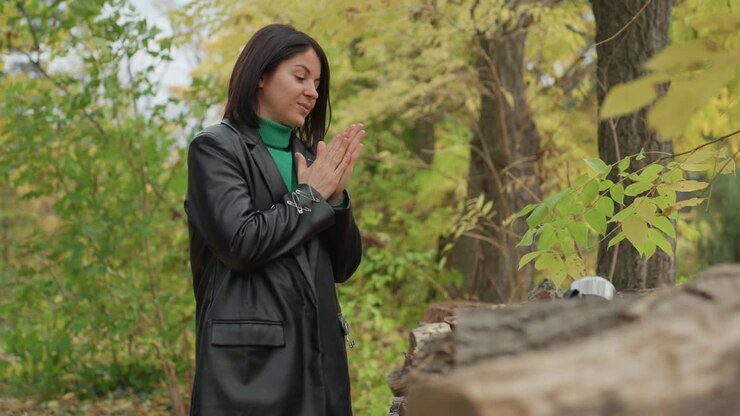 Woman in leather jacket warming hands beside log with thermos and mug, surrounded by autumn forest and golden foliage, green turtleneck pop of color, gentle smile and reflective mood, cozy rustic