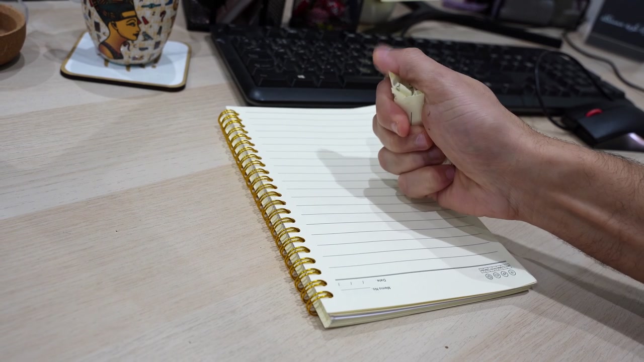 Close-up of a hand tearing handwritten notes from a spiral notebook on an office desk, with a keyboard, pen, and decorative mug in the background.