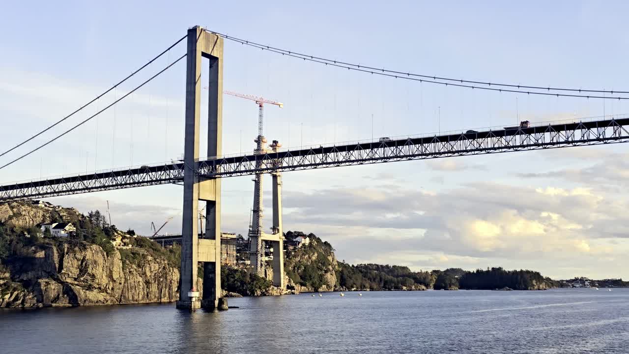 Camera moves forward from sea revealing old Sotra Bridge with traffic and new tower under construction