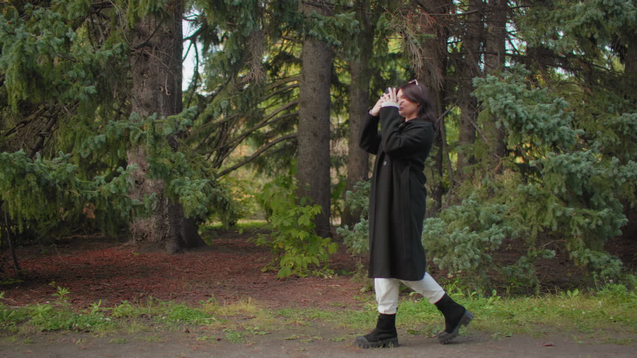 Signora elegante con cappotto autunnale che passeggia scattando foto tra i pini, vista laterale a media distanza, passeggiata lenta e tranquilla, telefono pronto, scena tranquilla in un parco naturale sotto la tenue luce del giorno