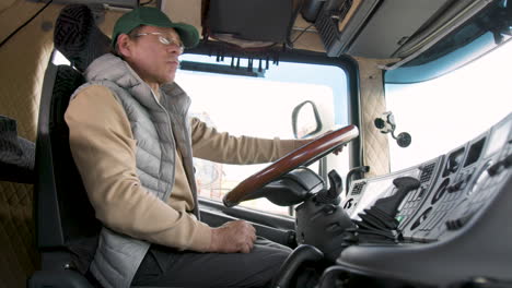 side view of caucasian older worker wearing cap and vest driving a truck in a logistics park