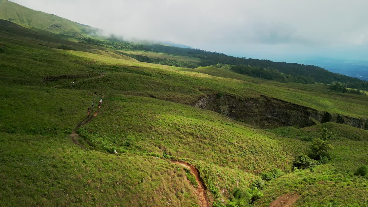 Hikers walk along a winding path over a green hill in Lombok under a partly cloudy but sunny sky, surrounded by lush grasslands and scenic views of nature in peaceful silence.