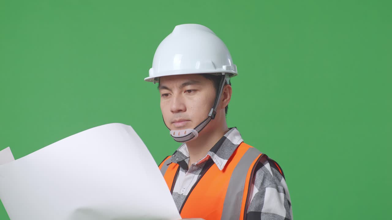 Close Up Side View Of Asian Male Engineer With Safety Helmet Looking At Blueprint In His Hands And Looking Around While Standing In The Green Screen Background Studio