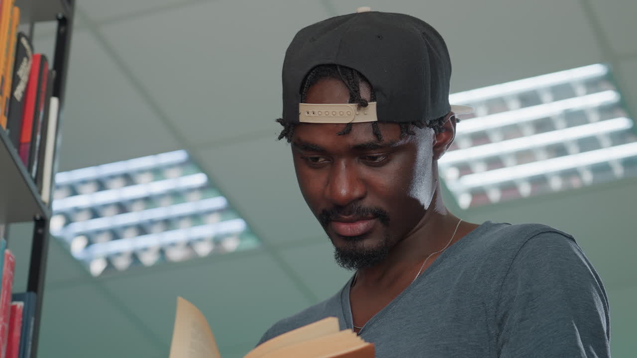Close up of young man in casual shirt and backwards cap reading hardcover book in library under bright fluorescent lights with focused expression, holding pages carefully