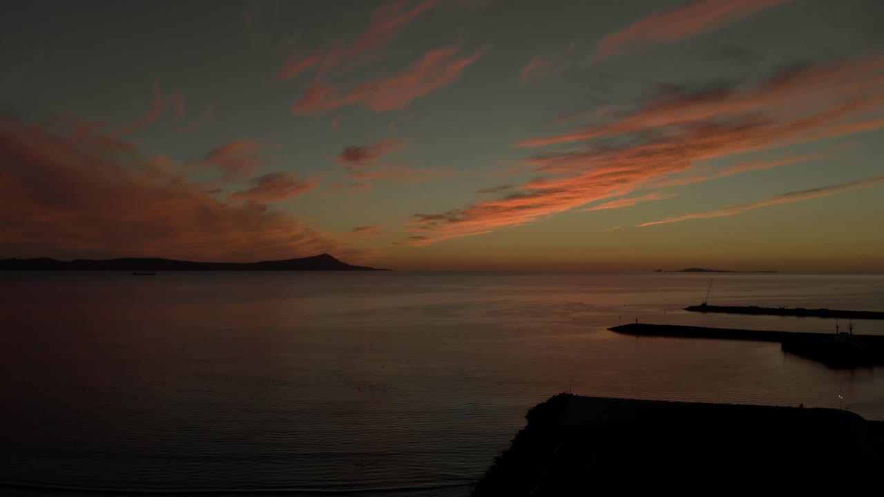 Aerial drone view of calm sea during sunset with vibrant sky and soft clouds, distant coastline and horizon creating peaceful seascape with warm evening light