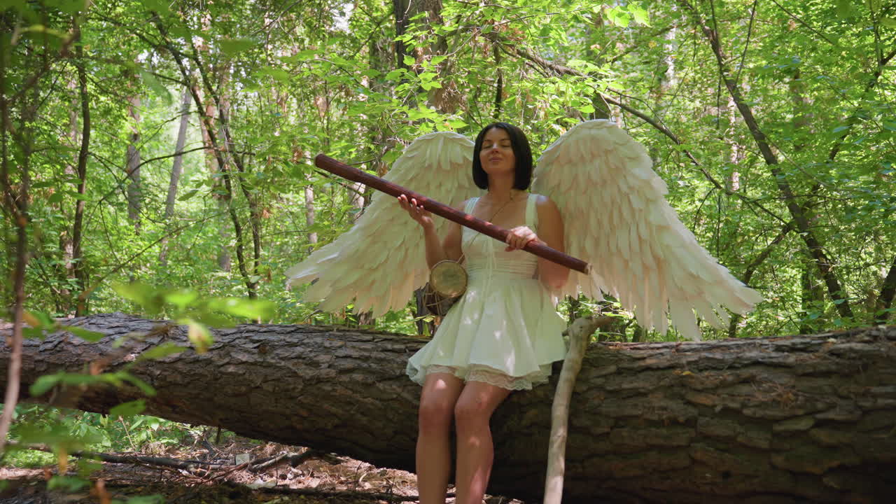 Fairy guardian woman with majestic white wings sitting on fallen tree in green forest holding long staff under gentle sunlight surrounded by lush foliage creating peaceful magical atmosphere