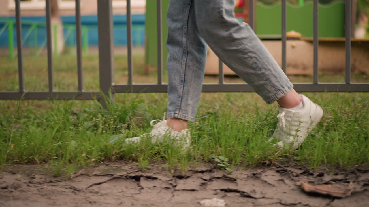 primer plano de los pies de la mujer en zapatillas blancas caminando a través de la hierba verde al lado de una valla de hierro, vaqueros de vaqueros enrollados ligeramente por encima de los tobillos, con vibrantes colores de patio de recreo borrosos en el fondo