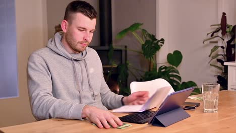 frustrated white young man meticulously examines bill on paper while seated at a table, disapprovingly shakes his head