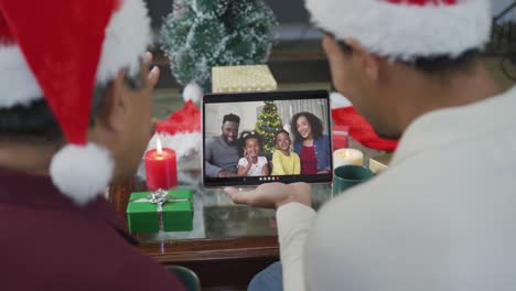 Padre-Birracial-Con-Hijo-Saludando-Y-Usando-Una-Tableta-Para-Una-Videollamada-Navideña-Con-La-Familia-En-La-Pantalla