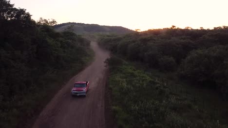 Classic-red-truck-drives-down-dusty-dirt-mountain-road-lined-with-trees,-aerial