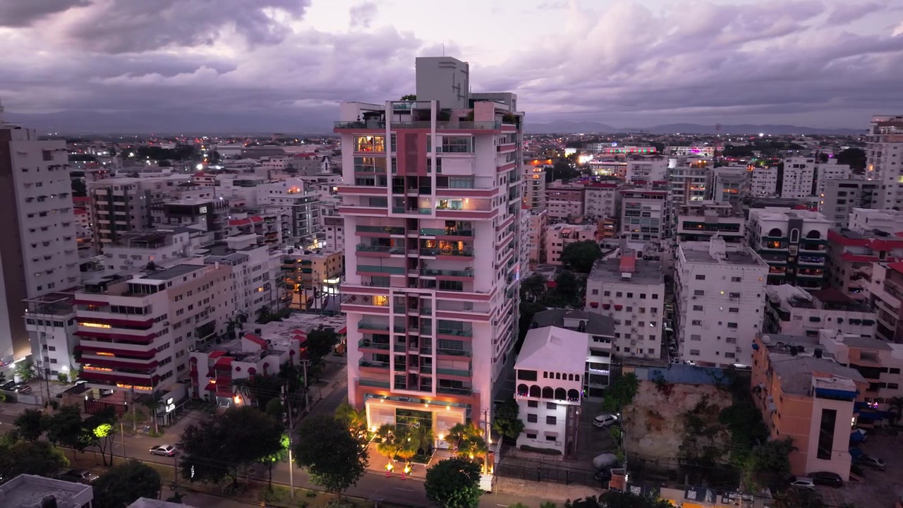 Toma aérea con dron con paneo lateral a través de un moderno edificio residencial de gran altura en medio del paisaje urbano iluminado de Santo Domingo al atardecer