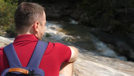 travel man with a backpack on his back sitting by mountain river enjoying nature