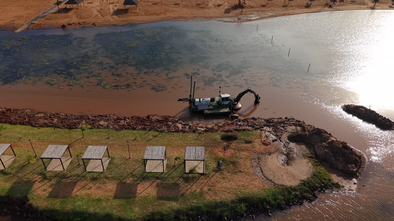 Amphibious dredging machine works along a muddy riverbank under the hot sun near Posadas, Misiones, Argentina, passing by rows of empty wooden shelters and calm water, drone panning shot