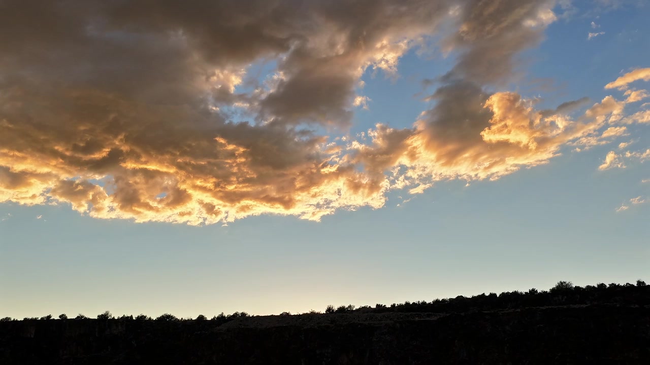 A 4k drone hyperlapse view of clouds shifting into vibrant gold colors during the sunset hour from below a dark silhouetted canyon ridge and tree-lined horizon.