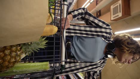 mujer sonriente con camisa a cuadros y auriculares en el cuello está caminando en la tienda de comestibles dirigiendo el carrito de compras con comida dentro