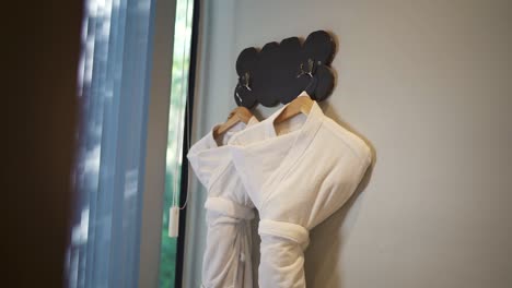 slow moving camera shot of white bath robes hanging on massage room in hotel, argentina. light grey wall and window on scene.