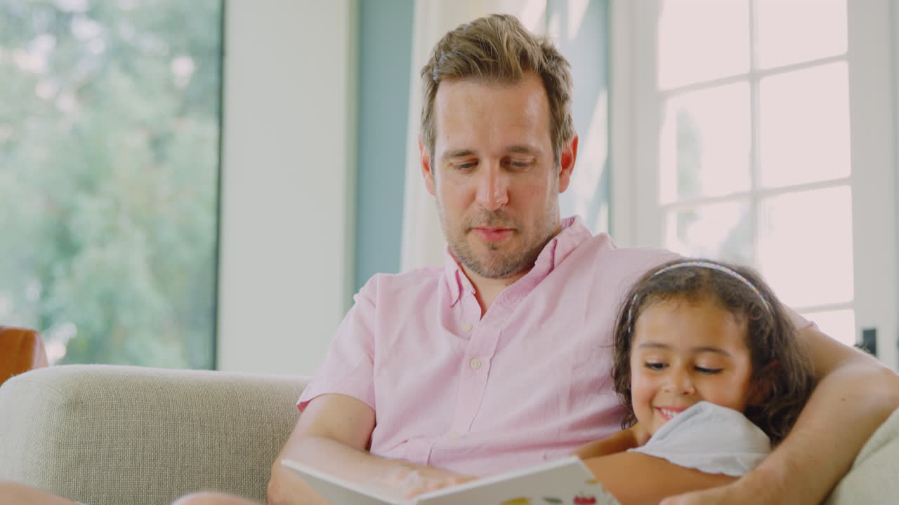padre e hija sentados en el sofá en casa leyendo un libro juntos