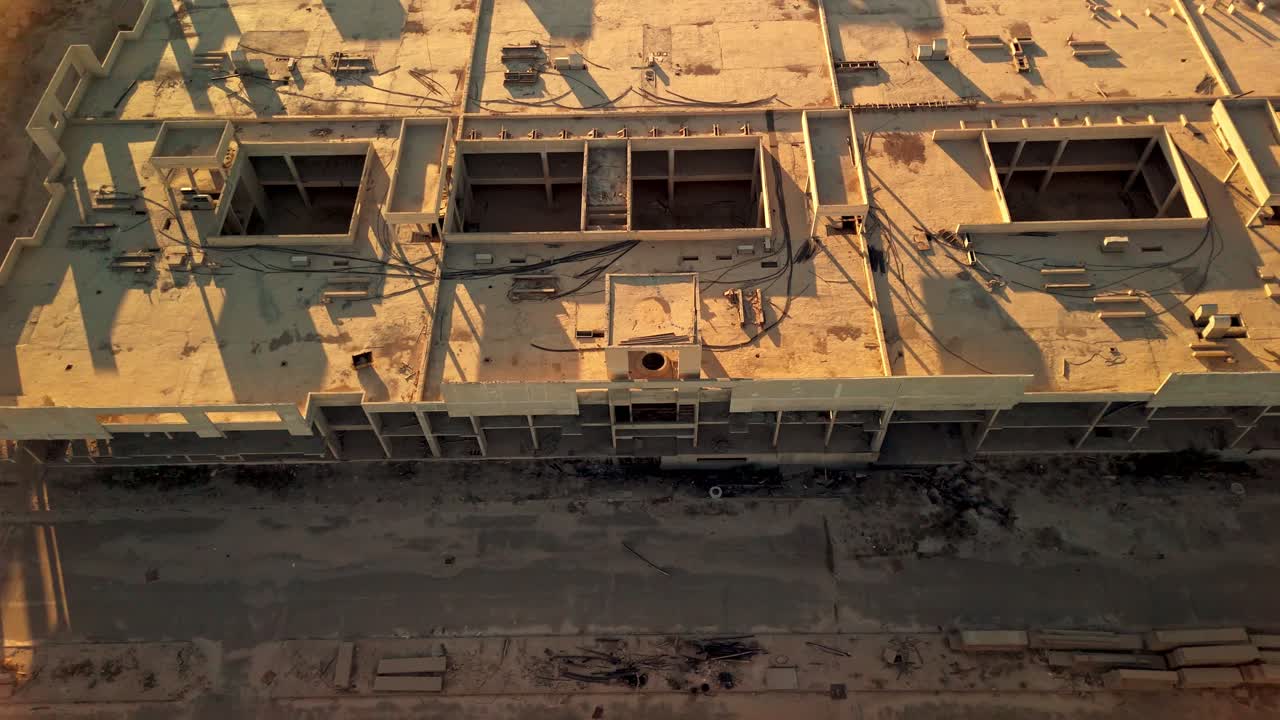 Ripresa aerea con drone 4K di un edificio in cemento incompiuto in un cantiere attivo durante l'ora d'oro. Lunghe ombre del tramonto su aperture sul tetto, strade sterrate e zona di lavoro