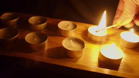 man lights tea candles in wooden tray in dark room as camera dollies