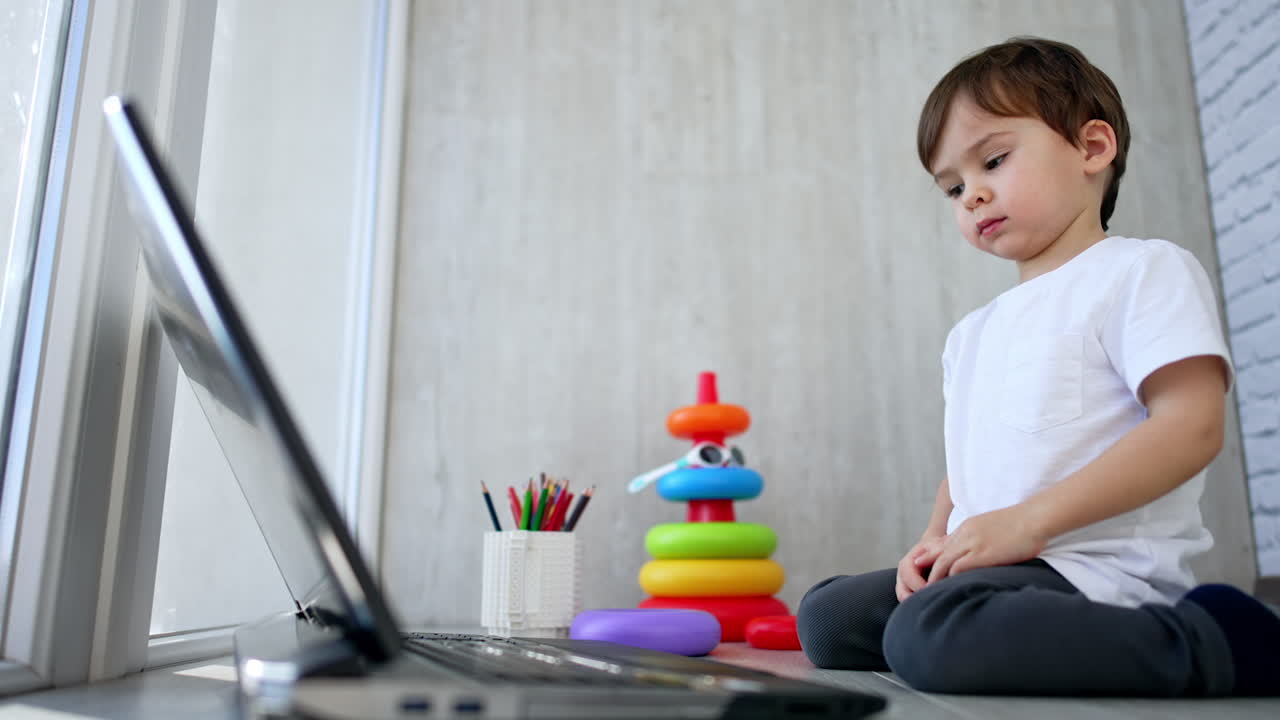 A cute little boy is sitting on the floor and watching cartoons on a laptop. The child is engaged in distance learning at home.