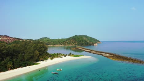 Rocky-barrier-protecting-calm-bay-of-turquoise-lagoon-with-calm-clear-water,-anchored-boats-on-shore-of-tropical-island,-Thailand