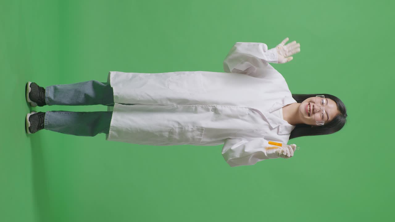 미소 짓는 아시아 여성 과학자의 몸은 실험관에 오렌지 액체와 함께 그녀의 손을 흔들고 실험실의 녹색 화면 배경에 서있는 동안 안을 말합니다.