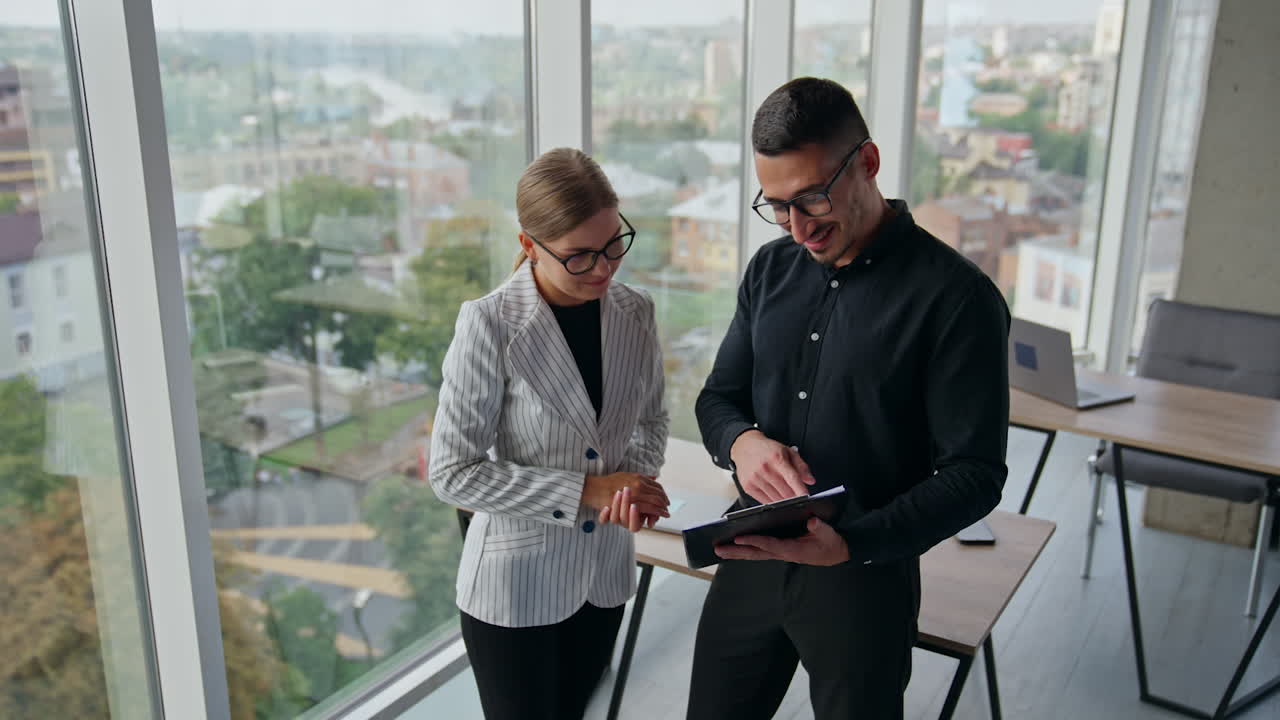 Company employees standing at office discussing papers. People talk, laugh looking at charts. Panoramic windows with cityscape at backdrop.