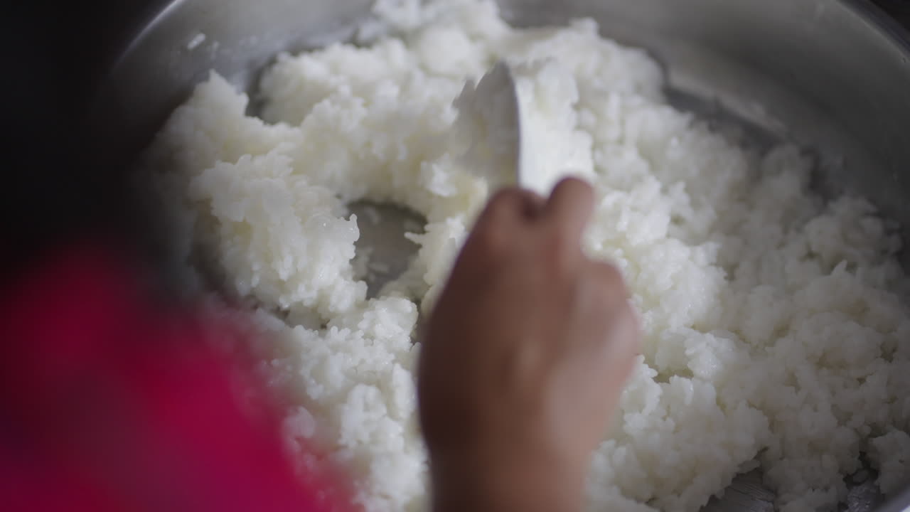 A person turns freshly cooked sushi rice in a pot, seen from an over-the-shoulder angle