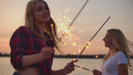 two girls are dancing with big bengal lights on the river coast. this is enjoyable summer evening on the open air party at pink sunset.