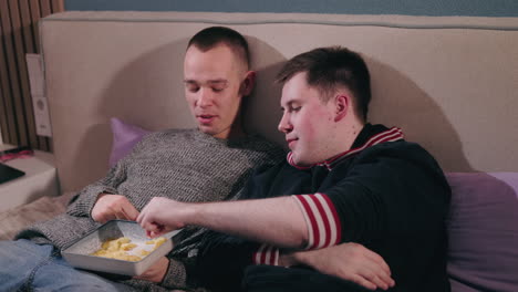 two men relaxing in bed, watching and eating chips