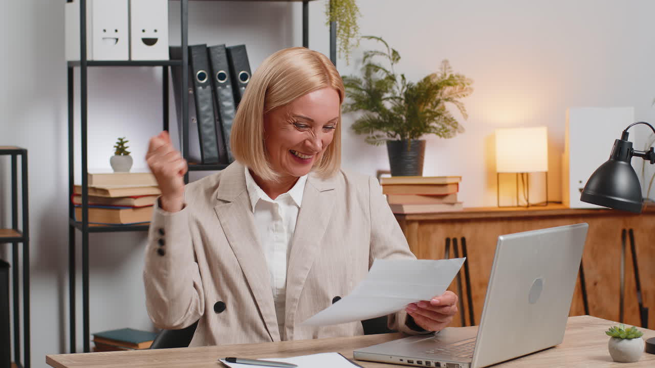 mujer de negocios madura exitosa con computadora portátil leyendo cartas y apretando el puño en el escritorio de la oficina en casa