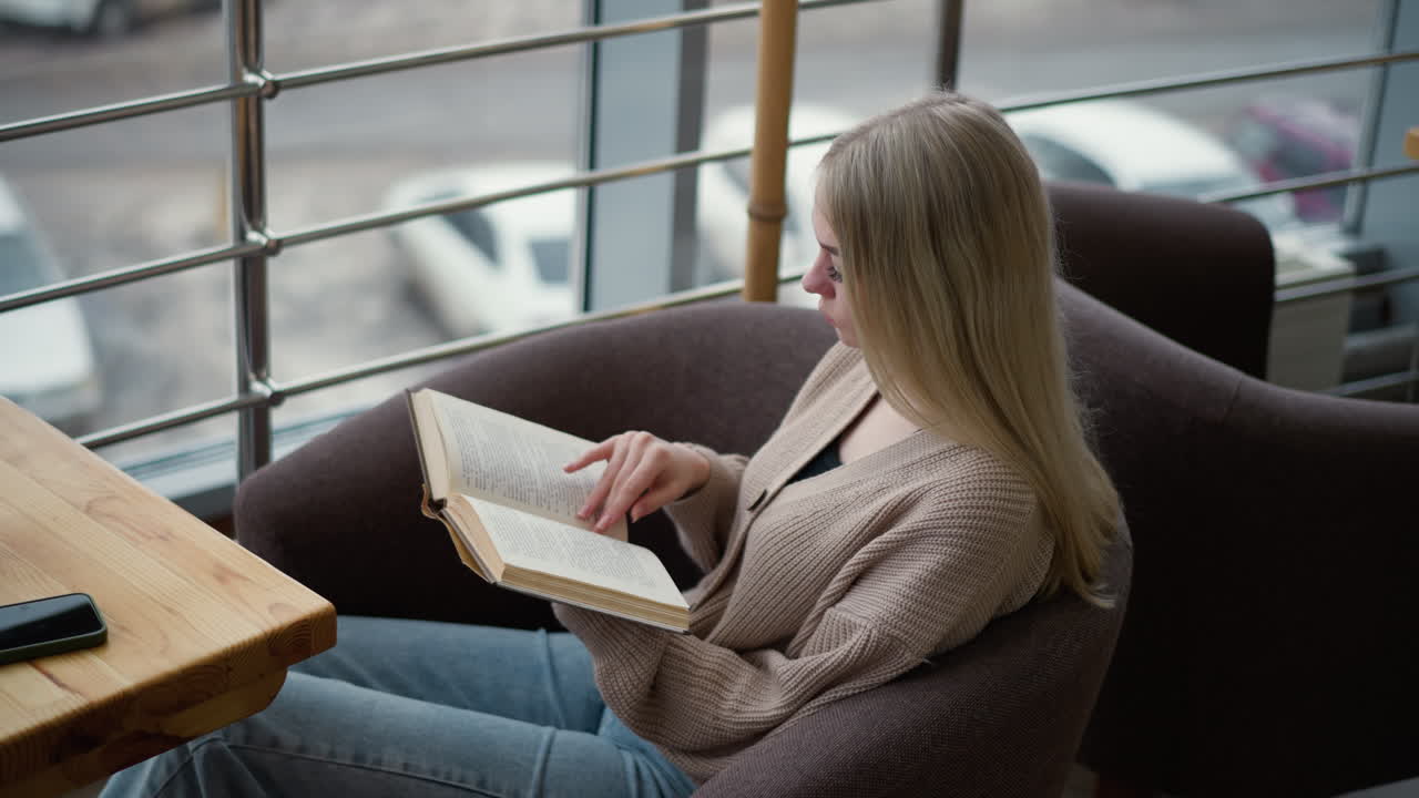 mujer sentada junto a la ventana leyendo un libro, volviendo a una nueva página con atención enfocada, inmersa en la literatura mientras la luz natural llena el espacio, creando una atmósfera de estudio pacífica