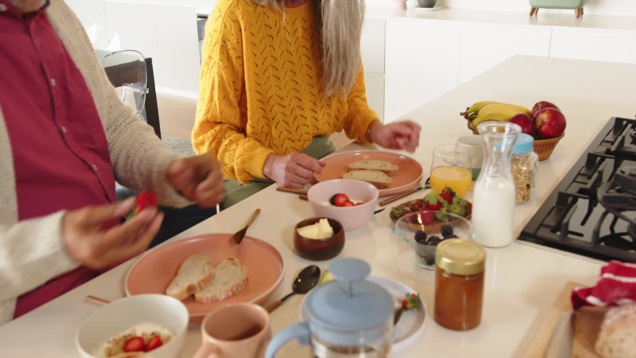Eating breakfast together, multiracial senior couple enjoying fresh fruit and coffee at home