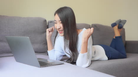 Glückliche-Asiatische-Junge-Frau,-Die-Zu-Hause-Auf-Dem-Sofa-Liegt-Und-Auf-Dem-Laptop-Tippt,-Erhält-Großartige-Nachrichten,-Die-Einen-Moment-Des-Sieges-Feiern,-Persönliche-Leistung,-Wurde-Eingestellt,-Stipendien-zulassungskonzept-Für-Die-High-School