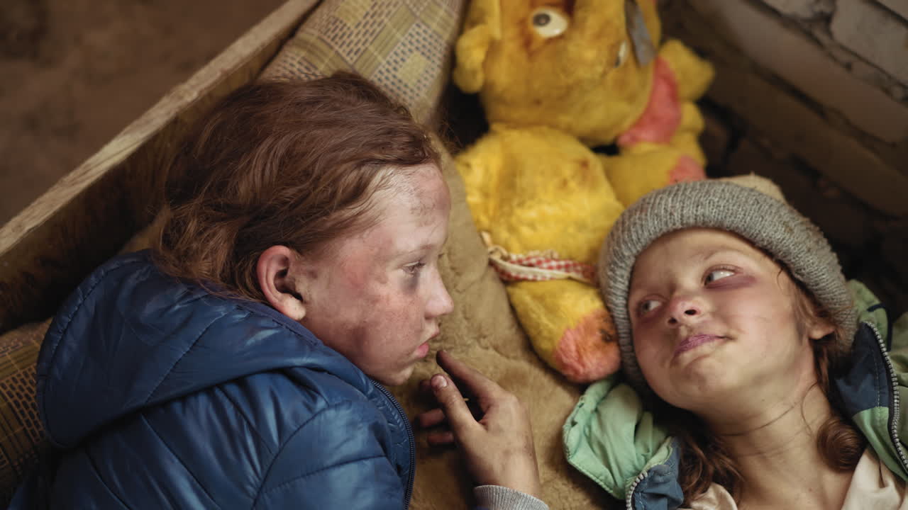 Two orphan children lie close on rough bedding beside worn yellow teddy bear inside damaged shelter, faces tired and dirty, sharing quiet connection and silent gaze