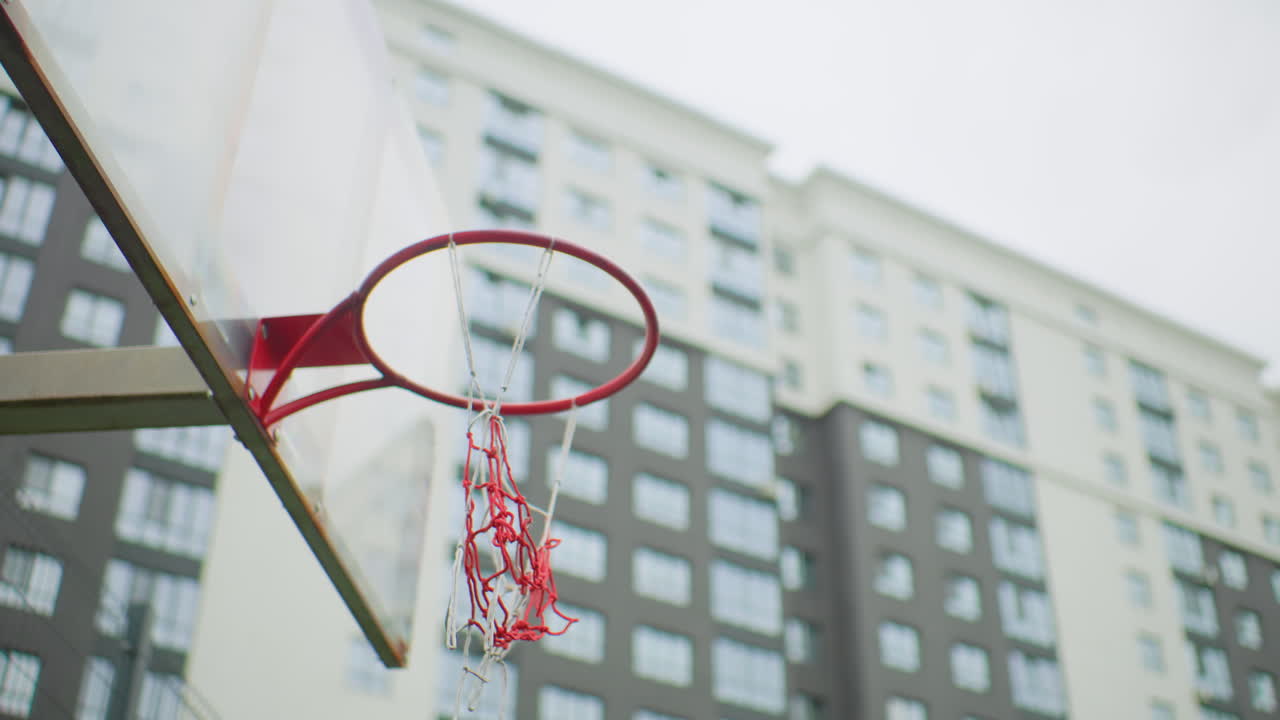 canestro da basket urbano con edificio cittadino sullo sfondo, traiettoria della palla verso il canestro, poi la rete si svuota, canestro in metallo rosso con rete a catena sfilacciata, cielo nuvoloso, finestre di grattacieli residenziali sfocate, tranquillo campo da gioco di quartiere