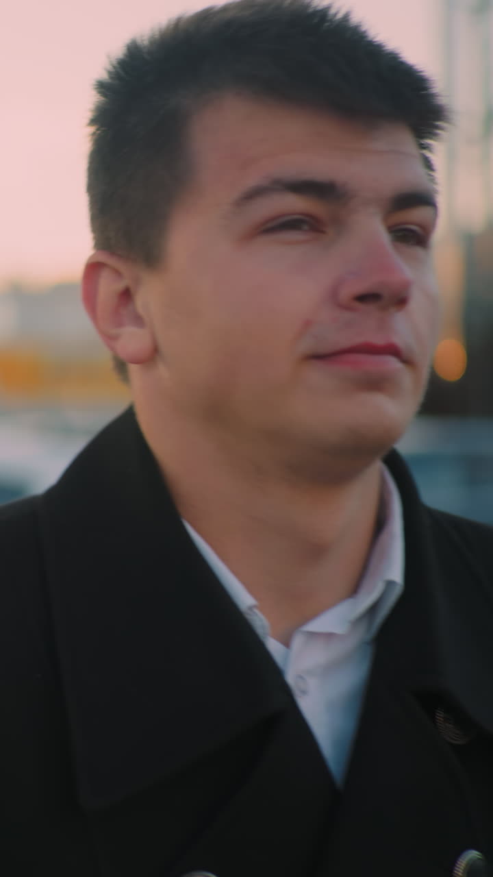 검은 코트를 입은 남자가 왼손에 브리프케이스를 들고 차 밖으로 나와, 배경에 주차된 차가 있는 건물을 향해 걸어가고, 하늘에 작은 은 구름, 황혼에 도시 설정