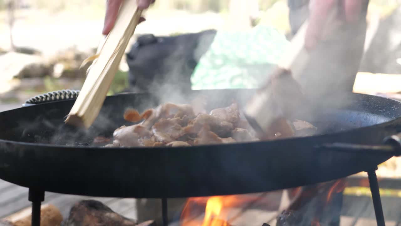 Close up shot of man stir-frying pieces of beef kebab meat in a muurikka griddle pan, over a hot wood fire in a camping environment