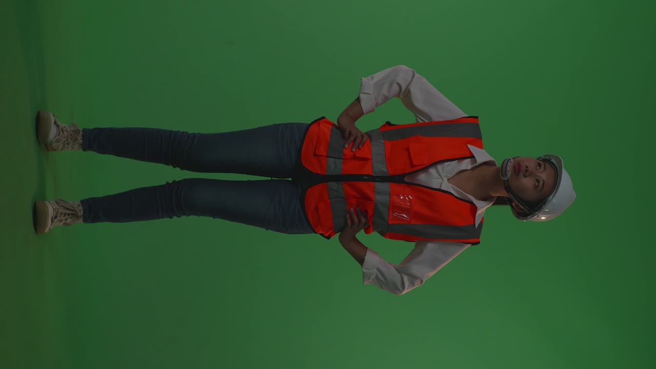 Full Body Of Asian Female Engineer Wearing Safety Helmet Looking Around While Standing With Arms Akimbo In The Green Screen Background Studio, During Sunset Or Sunrise Time