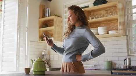 caucasian woman standing with hand on hip in cottage kitchen using smartphone, teapot on counter