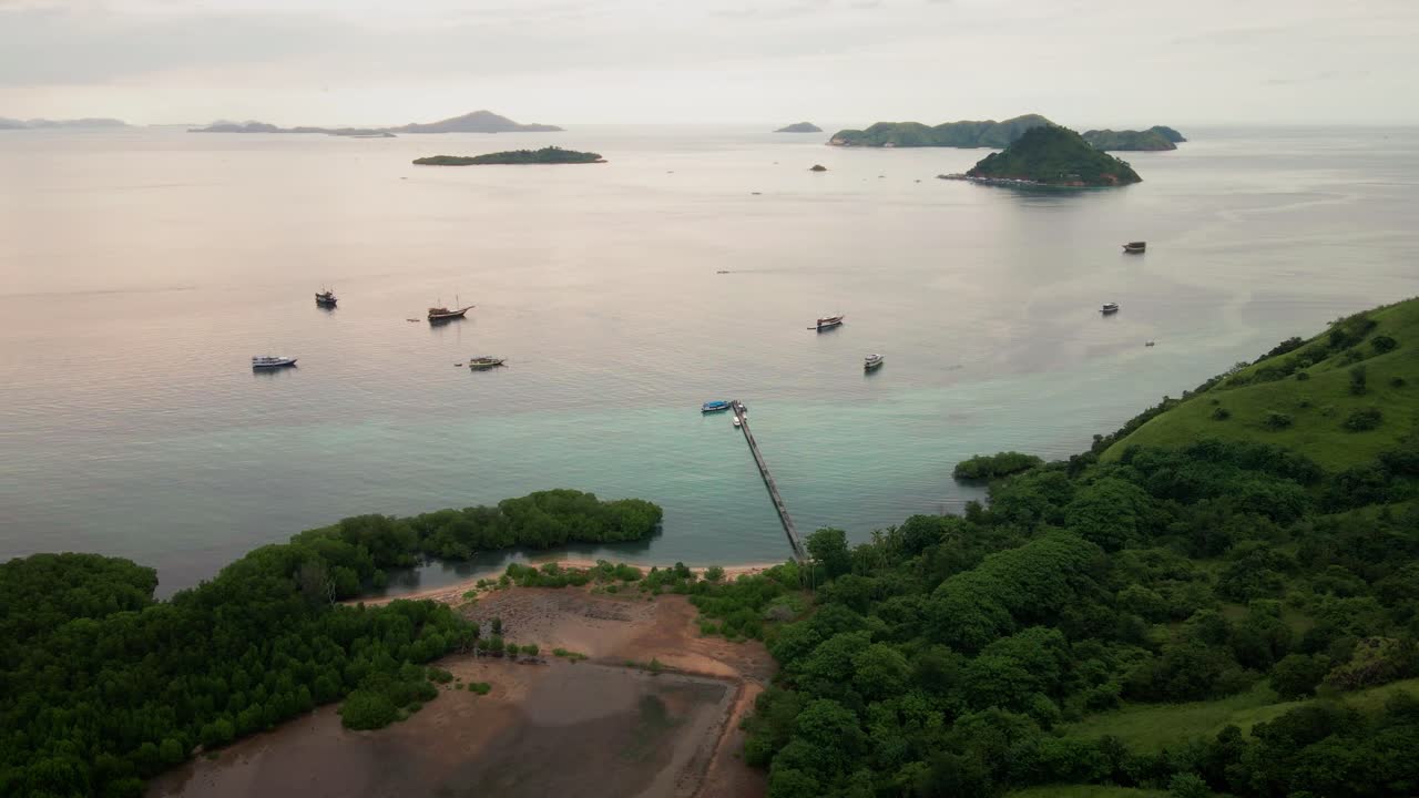A sunset view over the exotic tropical island of Komodo Island, peach-pink hues from the sunset reflect on the ocean—a romantic scene of green hills, clear waters, and boats gently sailing around