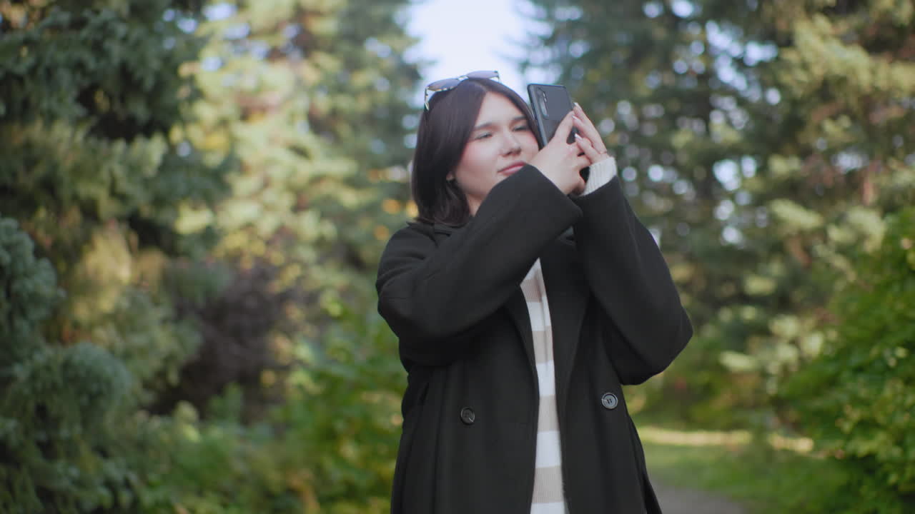 겨울 코트를 입은 우아한 여성이 숲길을 따라 걸어가는 하향 시점, 움직이는 다리와 청키한 부츠, 부드러운 햇빛, 푸른 나뭇잎, 사진 촬영 준비가 된 스마트폰, 평온한 산책 분위기