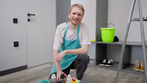 Portrait-of-a-confident-blond-man-in-a-white-T-shirt-as-a-cleaner-in-a-blue-apron-who-sits-on-the-floor-and-poses-near-cleaning-supplies-collected-in-a-gray-plastic-basin-and-while-cleaning-on-call-in-a-modern-apartment