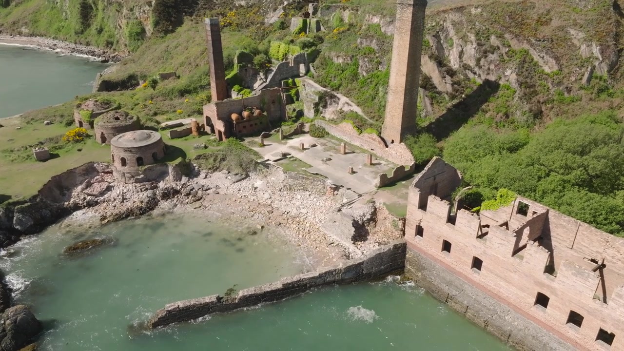 Ladrilleras de Porth Wen, Anglesey, Gales. Órbita lenta mostrando edificios dilapidados y abandonados, hornos y chimeneas con agua de mar tranquila y clara lamiendo la orilla en un día soleado.