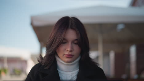 young woman seated in an outdoor setting with wind gently swaying her hair, deep in thought, she is dressed in a white turtleneck and black coat with blur background