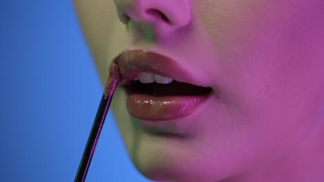 Extreme-close-up-of-a-young-pretty-woman-applying-shiny-lip-gloss-on-her-beautiful-red-lips-while-getting-ready-to-go-out-with-friends-for-club-night-or-date-with-purple-contrast-light-in-her-face