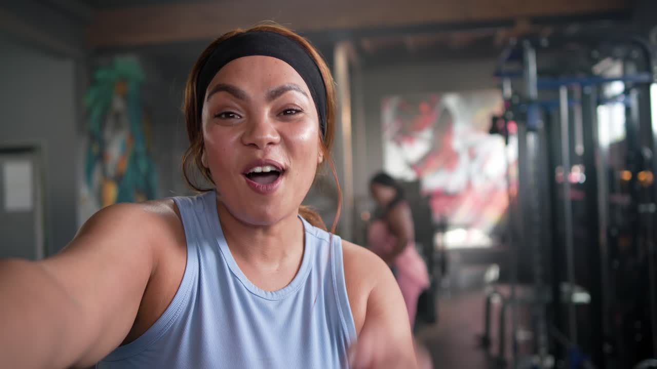 Sportliche Partnerinnen in Activewear, Frontfrau erreicht Kamera, Vlogging im Fitnessstudio, zeigt auf Hanteln